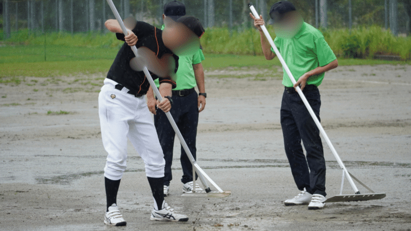 雨の中、水がたまったマウンド周りをトンボでならす監督と審判